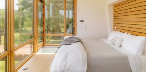 Bright modern bedroom with a neatly made bed, light wood accents, and large glass doors opening to a deck with views of trees.