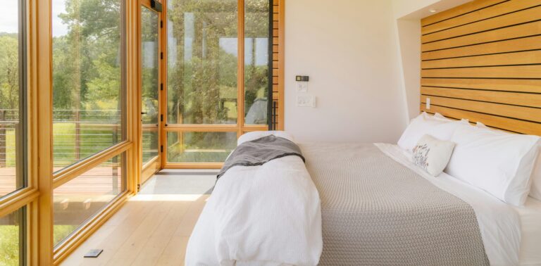 Bright modern bedroom with a neatly made bed, light wood accents, and large glass doors opening to a deck with views of trees.