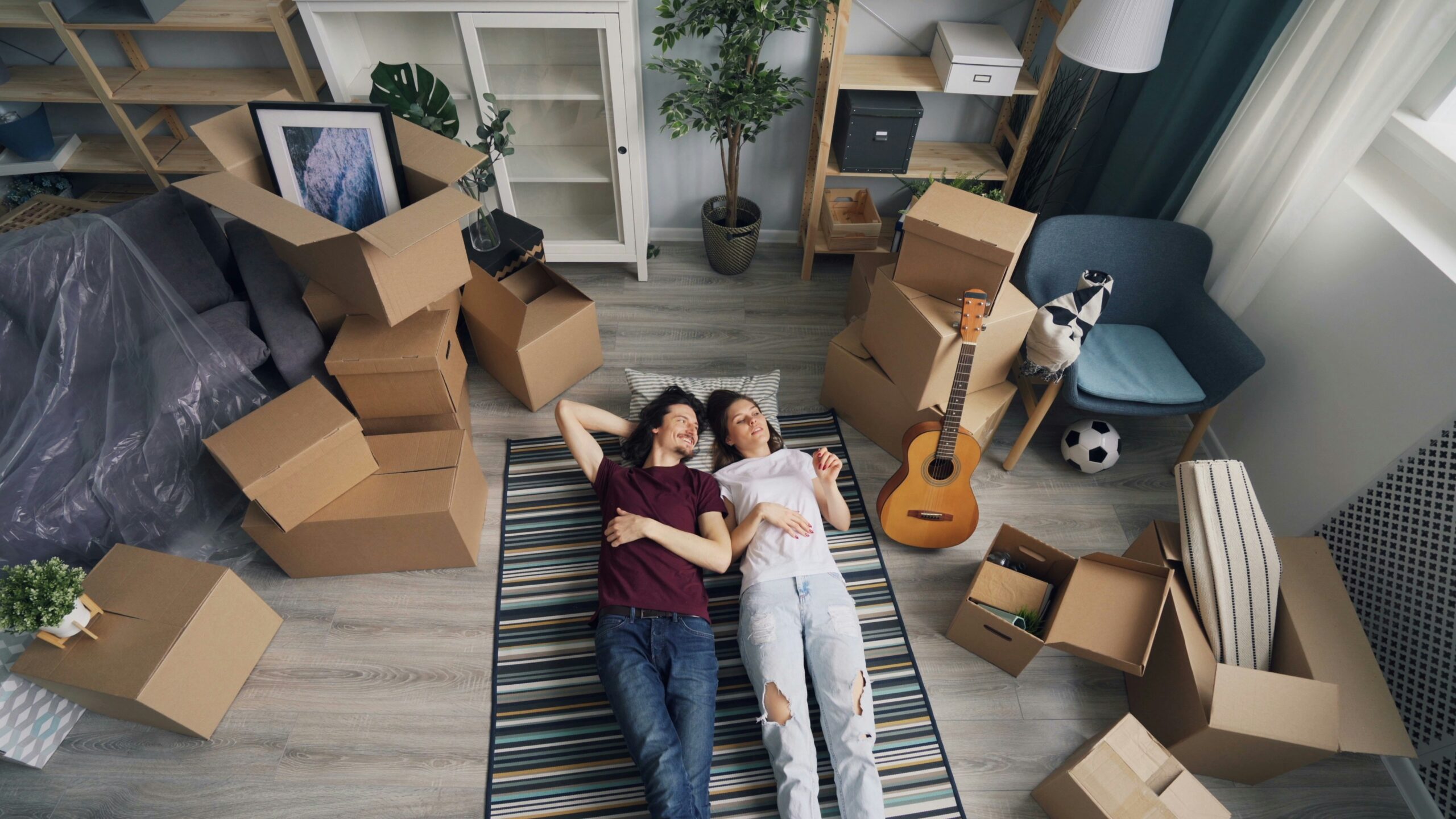 Two people lying on a striped rug in a living room surrounded by cardboard moving boxes, furniture, shelves, plants, a guitar, and household items.