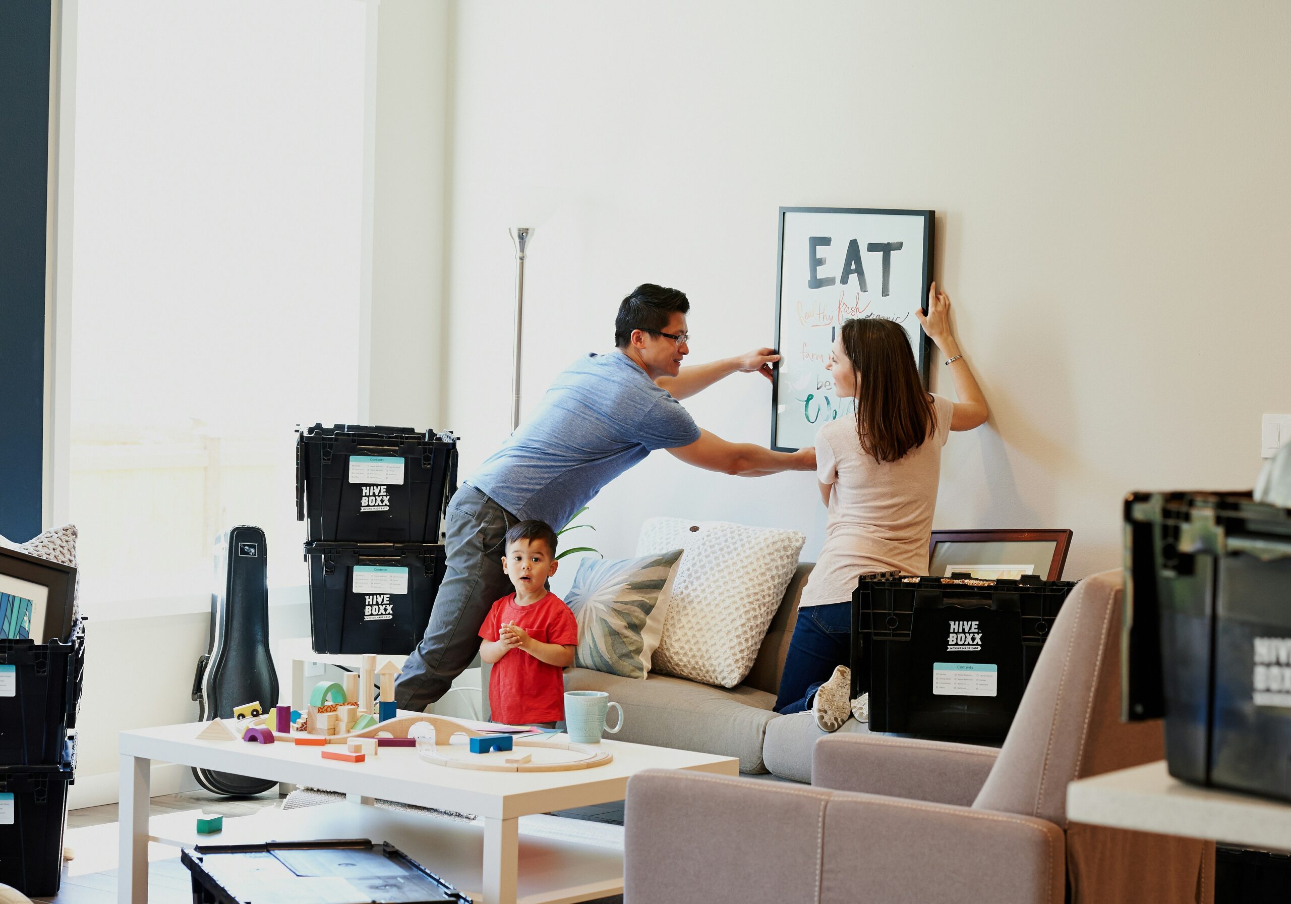 Three people in a living room unpacking after a move, with two people hanging framed wall art above a sofa while a child stands nearby, surrounded by labeled moving boxes and household items.