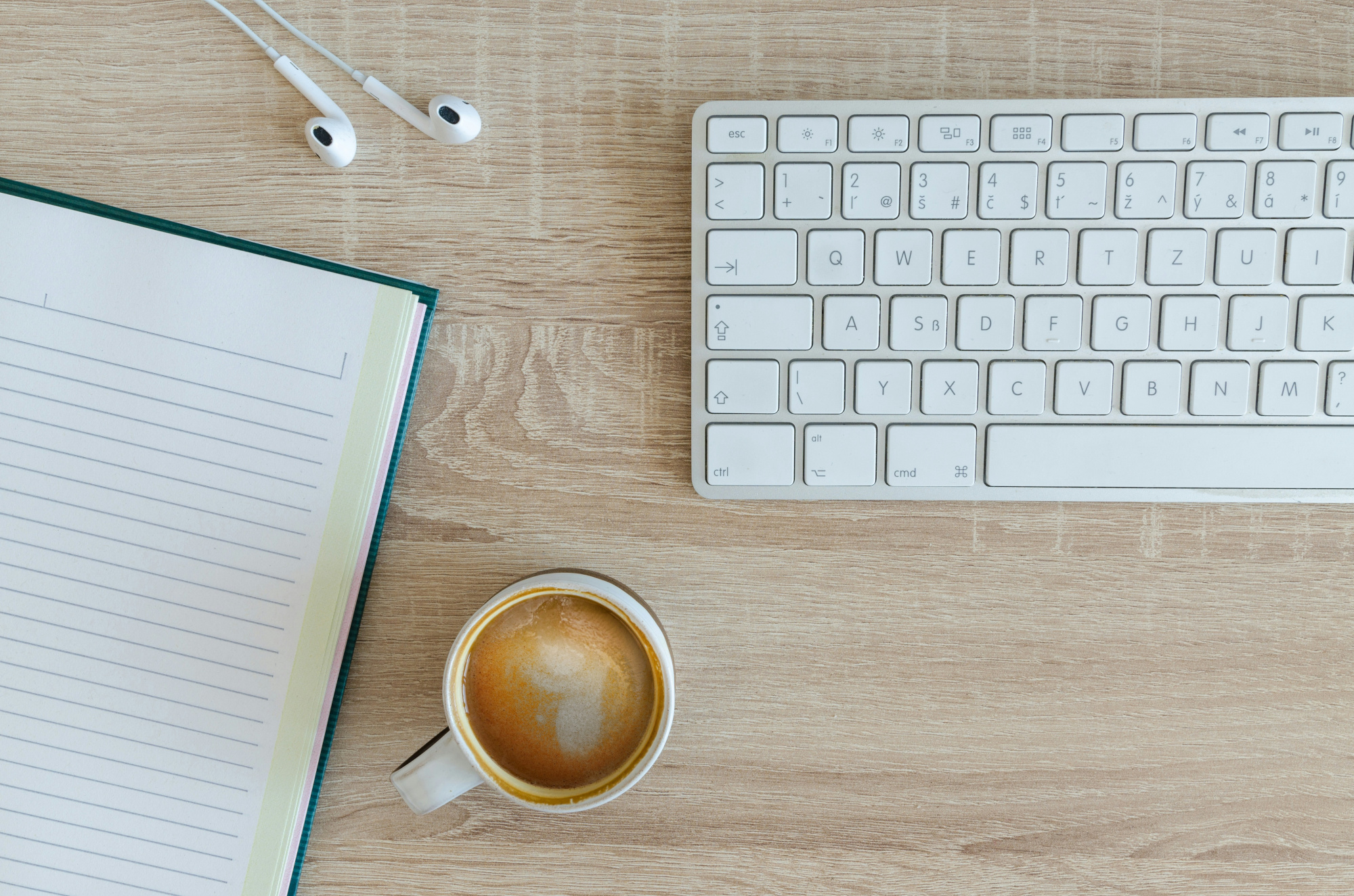 Top-down view of a minimalist workspace with a white keyboard, earbuds, notebook, and a cup of coffee on a wooden desk