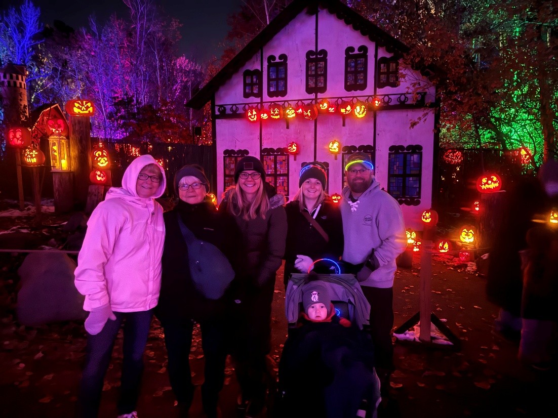 Group gathered at a Jack-O-Lantern Spectacular at night, surrounded by glowing carved pumpkins, colorful illuminated trees, and a Halloween-themed building display.